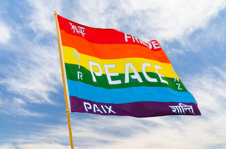 Waving rainbow varicolored peace flag with multilingual pacific text flying on wind against blue skyの写真素材