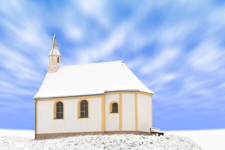 Standing alone detached small building of winter snow-covered Catholic chapel against blue cloudy skyの写真素材