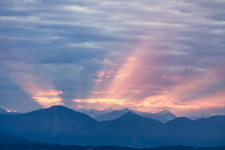 First rays of the rising sun on dawning cloudy sky over dark blue morning Alps mountainsの写真素材