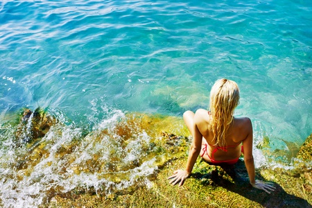 Young attractive woman sitting on the rocks near the blue water of the sea and taking sunbath in a beautiful day of summerの写真素材