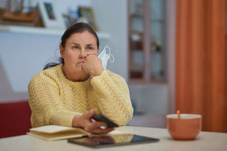 Sad senior woman in yellow sweater with mask and smart phoneの写真素材