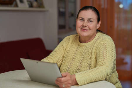 Senior woman in yellow sweater looking in the camera with tabletの写真素材