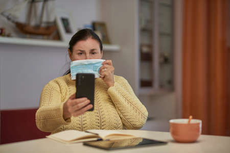 Senior woman in yellow sweater looking at smartphone with maskの写真素材