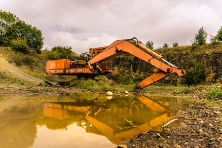 abandoned orange excavator reflecting in muddy water of puddleの写真素材