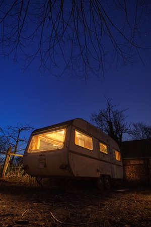 Old white caravan trailer with yellow illumination in backyard at stary blue night