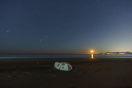 Sandy sea beach with white boat and illuminated Amalfi Coast at night with Orion and Pleiades constellation and moon rising, Paestum, Campania, Italyの写真素材