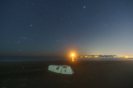 Beach with white boat and illuminated coast at hazy night with Orion and Pleiades constellation and moon rising, Paestum, Campania, Italyの写真素材
