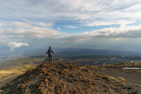 Hiking person looking at view of volcanic landscape of Mount Etna with black lava and yellow grass on partly cloudy winter day, Catania, Sicily, Italyの写真素材