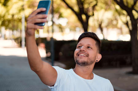 young, handsome, Hispanic Latin boy with short hair, beard and white flannel is taking a picture of himself with his phone and is in a park on a sunny day.の写真素材