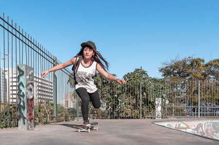 pretty young white hispanic latin girl in black clothes and gray cap sitting on a skate on the floor...