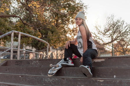 pretty young white hispanic latin girl in black clothes and gray cap sitting on a skate on the floor...