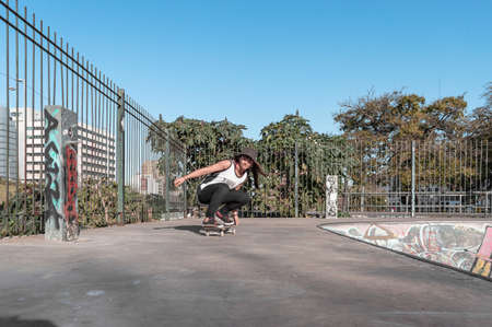 pretty young white hispanic latin girl in black clothes and gray cap sitting on a skate on the floor of a skate park with blue sky and trees in the background using her phone. technology concept.の写真素材