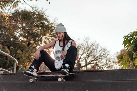 pretty young white hispanic latin girl in black clothes and gray cap sitting on a skate on the floor...