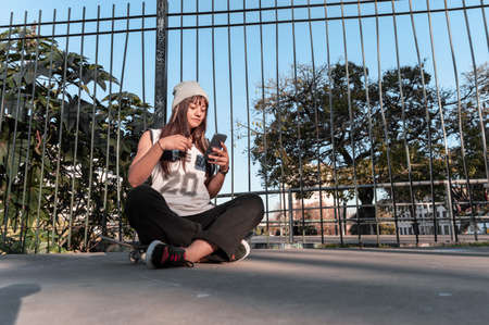 pretty young white hispanic latin girl in black clothes and gray cap sitting on a skate on the floor...