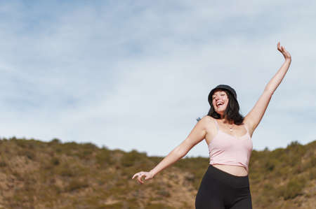 horizontal portrait of a pretty young hispanic latin girl white in pink posing with her hands on her black hat looking to the side with an astonished face staring at a valley with arid vegetationの写真素材