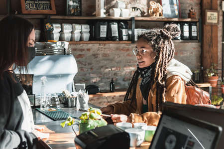 pretty young white hispanic latin girl with glasses, dreadlocks, brown jacket and backpack and black scarf smiling with the menu in her hands ordering a espresso coffee at the counter of a coffee shopの写真素材