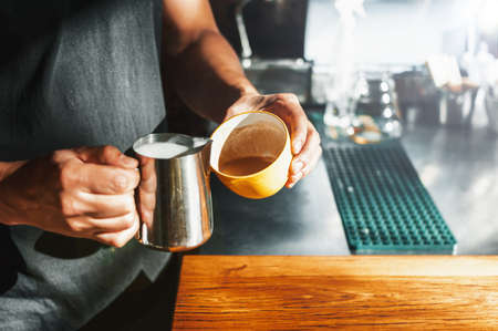 brunet hispanic latin adult male waiter, dressed in black, holding a yellow mug and a stainless steel carafe, preparing a latte at the bar of a nice coffee shop.の写真素材