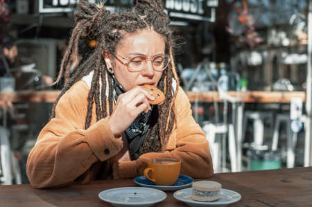 pretty young white hispanic latin girl with black bandanna, brown corduroy jacket, glasses and dreadlocks sitting outside a coffee shop with her mouth open eats a chocolate chip cookie.の写真素材