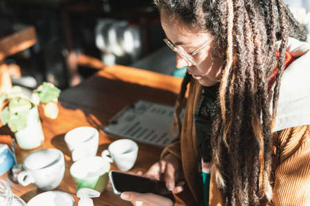 pretty young white hispanic latin girl with glasses, brown corduroy jacket, black scarf and dreadlocks at the bar of a nice cafeteria checking her cell phone, restaurant concept.の写真素材