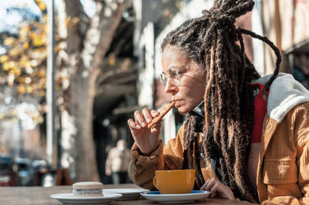 beautiful young white hispanic latin girl wearing a brown corduroy jacket, black bandanna, glasses and dreadlocks, sitting at a wooden table outside a coffee shop eating a chocolate chip cookie.の写真素材