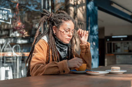 pretty white hispanic latin girl with dreadlocks, brown corduroy jacket, black bandana and glasses, is with her eyes closed enjoying a coffee and cookies sitting outside the coffee shop on a sunny dayの写真素材