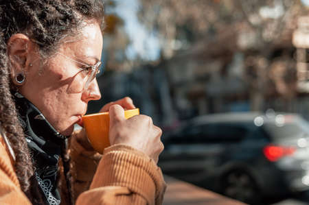 close up side shot of pretty young white hispanic latin girl with dreadlocks, glasses, black bandanna and brown corduroy jacket, drinking coffee from a yellow cup outside the coffee shop.の写真素材