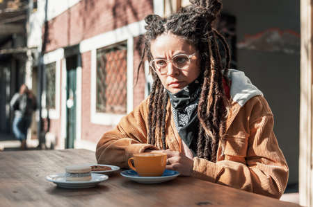 pretty young white hispanic latin girl with glasses, black scarf, brown corduroy jacket and dreadlocks enjoys sitting looking away and thinking at a coffee table on the street with a cup of coffee.の写真素材