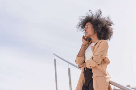 pretty young black Hispanic Latina college girl with afro, glasses and yellow jacket, talking on the phone and holding a blue folder with the sky in the background with copy spaceの写真素材
