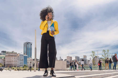 pretty young black Hispanic Latina college girl with afro, glasses and yellow jacket, talking on the...