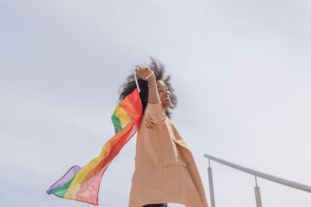 pretty young brunette latina girl with afro, happy, smiling and dancing contentedly waving a gay pride flag with the sky in the background outdoors on a sunny day.の写真素材