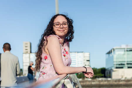 beautiful young Latina hispanic girl, white, with curls and glasses, stands on a pier, leaning on a...