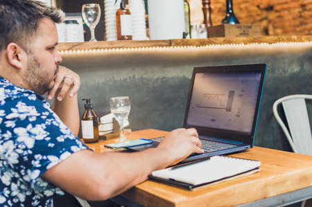 Young Hispanic Latino man with his computer and notepad, analyzing trading charts sitting in a...