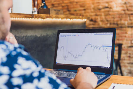 man watching laptop screen with financial charts and trading graphs, with one hand on keyboard, sitting inside restaurant, working as trader.の写真素材