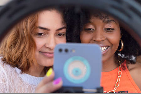 two multiethnic young hispanic latina girl friends, happy and smiling, checking social networks and surfing the internet on a phone on a tripod with a light ring. lifestyle and technology concept.の写真素材