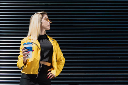 beautiful young blonde hispanic latin girl, dressed in black, jacket and yellow glasses, posing with a reusable coffee cup, fashion and lifestyle concept.の写真素材