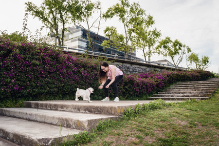 pretty young white hispanic latina girl, with lenses and curls, smiling contentedly in the park playing with her white poodle dog on the stairs next to a nice purple flower garden, lifestyle concept.の写真素材