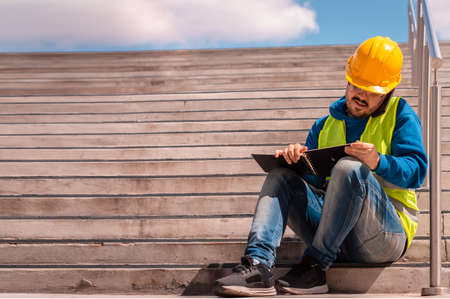 latino worker with yellow hard hat, glasses and green vest, sitting on a staircase with a diary in his hands, talking on the phone.の写真素材