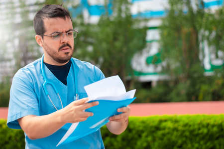 young Latin doctor, wearing glasses, outside the hospital, reading a medical chart and holding a...