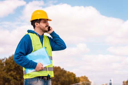 Latin worker with yellow hard hat, glasses and green vest, talking on the phone and holding a folder...