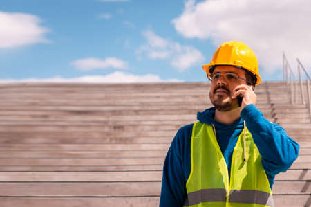latin worker with yellow hard hat, glasses and green vest, on a staircase talking on the phone