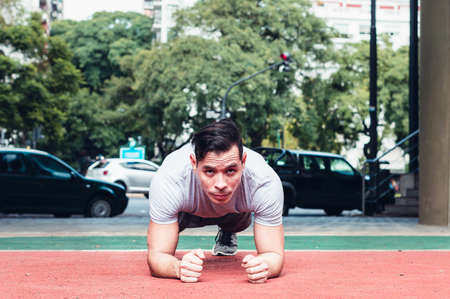 young athletic latin guy, white skin, doing push-ups, dressed in gray clothes and in a fitness plaza with an urban environment, outdoors.の写真素材