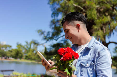 young caucasian man in love with flowers in hand sending message using his phone outdoors in a public park with natural habitat a lagoon and trees in the backgroundの写真素材