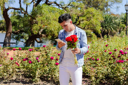 caucasian man standing in the garden of a park with many flowers and trees behind him, with a bouquet of flowers in his hand worried reading messages from his girlfriend on his phone. Valentine's Day.の写真素材