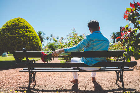 rear view of young caucasian hispanic latin man with a bouquet of flowers in his hand, sitting on a wooden bench in a beautiful garden.の写真素材