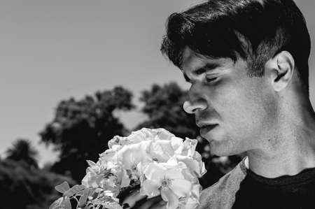 dramatic and romantic young caucasian argentinian man in black and white with eyes closed smelling a flower in the park, with the sky on background and copy space.の写真素材