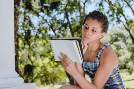 beautiful young brunette latin argentinian girl is sitting with a notebook and a pen in a park, with...
