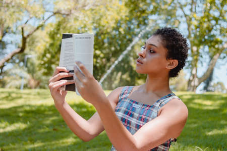 strong young brunette hispanic latin girl with short hair holding a book and reading outside in a public park with trees and nature around, lifestyle concept.の写真素材