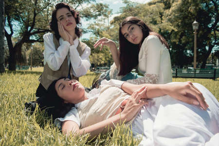 three sweet retro latin argentinian girls sitting on the grass resting in a public park with trees...