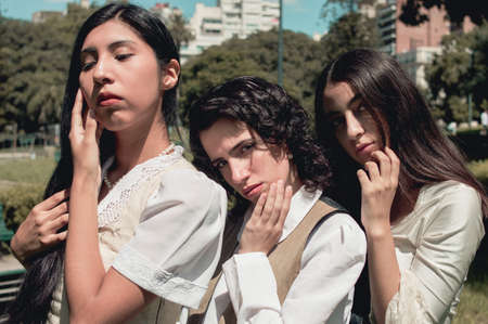 three pretty young hispanic latina argentinian girls standing in a park posing touching their faces,...
