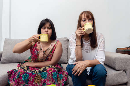 front view of young woman with down syndrome sitting having a cup of coffee with her mom in the living room, enjoying watching a movie together. family and inclusion conceptの写真素材
